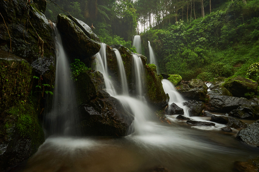 View of Air Terjun Grenjengan Kembar cascading over dark mossy rocks surrounded by lush green forest in Kecamatan Pakis Jawa Tengah Indonesia.
