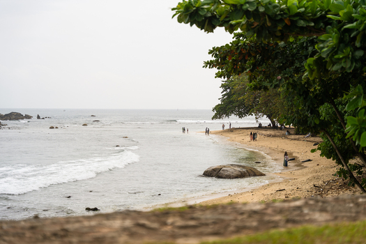 View of people walking along a sandy beach with lush green trees and a large rock in the shallow water under an overcast sky in Galle Southern Province Sri Lanka.