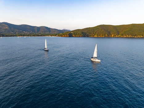 Aerial view of two sailboats sailing on the calm blue waters of Lacona Bay with green hills in the background in Lacona Tuscany Italy.