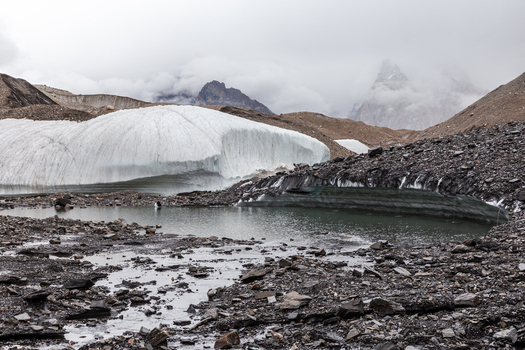 View of the Baltoro Glacier with a meltwater pool surrounded by rocky moraine and distant mountain peaks under a cloudy sky in Baltoro Gilgit Baltistan Pakistan.