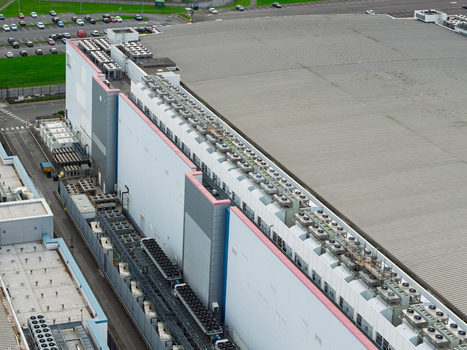 Aerial view of a large data center facility featuring numerous industrial cooling units on its expansive roof adjacent parking areas and paved grounds in the United Kingdom.
