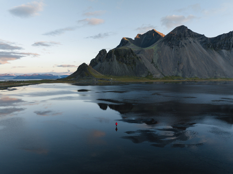 Aerial view of a lone figure in red stands on the black sand beach reflecting the majestic Vestrahorn mountain under the soft glow of dawn Höfn Iceland. Aerial view of a lone figure in red stands on the black sand beach reflecting the majestic Vestrahorn mountain under the soft glow of dawn Höfn Iceland.