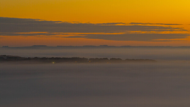 Aerial view of a landscape blanketed in thick morning fog with a line of trees emerging above the mist under a vibrant orange sunset sky in Salisbury England United Kingdom. Aerial view of a landscape blanketed in thick morning fog with a line of trees emerging above the mist under a vibrant orange sunset sky in Salisbury England United Kingdom.