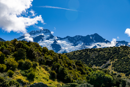 View of snow capped mountains rising majestically behind a foreground of lush green hills under a bright blue sky with scattered clouds Mount Cook Canterbury Region New Zealand. View of snow capped mountains rising majestically behind a foreground of lush green hills under a bright blue sky with scattered clouds Mount Cook Canterbury Region New Zealand.