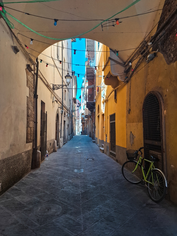 Pisa Tuscany Italy 27 April 2026 View of a narrow historic street in Pisa with yellow buildings cobblestone pavement and a bicycle parked against a wall under string lights. Pisa Tuscany Italy 27 April 2026 View of a narrow historic street in Pisa with yellow buildings cobblestone pavement and a bicycle parked against a wall under string lights.