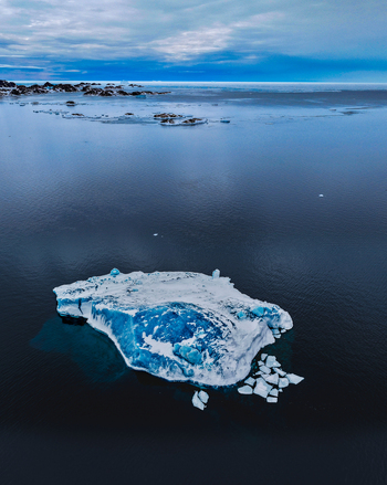 Aerial view of a large blue iceberg floating in the dark arctic waters under a cloudy sky near the coast in Tasiilaq Sermersooq Municipality Greenland. Aerial view of a large blue iceberg floating in the dark arctic waters under a cloudy sky near the coast in Tasiilaq Sermersooq Municipality Greenland.