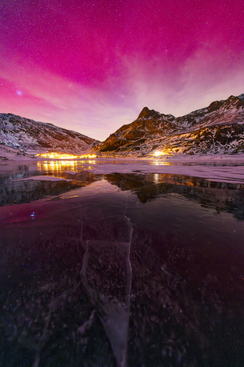 View of a frozen lake with ice cracks reflecting a vibrant pink night sky and snowy mountain peaks in Madesimo Lombardy Italy. View of a frozen lake with ice cracks reflecting a vibrant pink night sky and snowy mountain peaks in Madesimo Lombardy Italy.