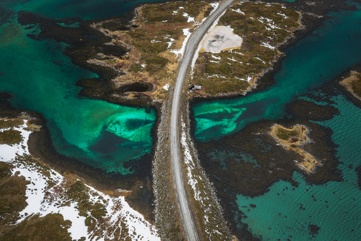 Aerial view of a road cutting through the vibrant turquoise waters and rocky islands where snow dusted land meets the sea Flakstadbruene Nordland Norway. Aerial view of a road cutting through the vibrant turquoise waters and rocky islands where snow dusted land meets the sea Flakstadbruene Nordland Norway.