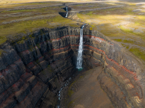 Aerial view of cascading waterfall plunges from layered cliffs into a rugged gorge contrasting starkly with the flat grassy plateau above Egilsstaðir Iceland. Aerial view of cascading waterfall plunges from layered cliffs into a rugged gorge contrasting starkly with the flat grassy plateau above Egilsstaðir Iceland.