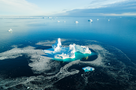 Aerial view of a large jagged iceberg floating in the deep blue waters of the Arctic Ocean surrounded by scattered sea ice and calm clear water in Greenland. Aerial view of a large jagged iceberg floating in the deep blue waters of the Arctic Ocean surrounded by scattered sea ice and calm clear water in Greenland.