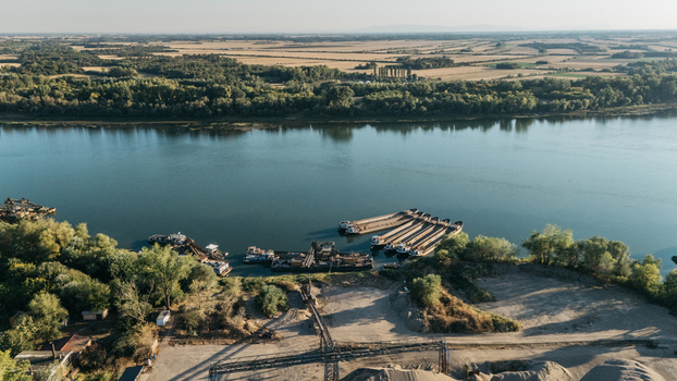 Aerial view of barges moored on the Sava River near an industrial site with gravel piles and lush green trees in Sremska Mitrovica Vojvodina Serbia.