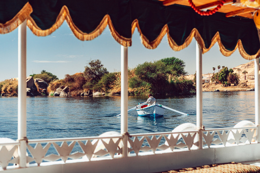 View of a person rowing a small boat on the Nile River seen from a traditional vessel with decorative curtains and lush islands in the background Aswan Aswan Governorate Egypt. View of a person rowing a small boat on the Nile River seen from a traditional vessel with decorative curtains and lush islands in the background Aswan Aswan Governorate Egypt.