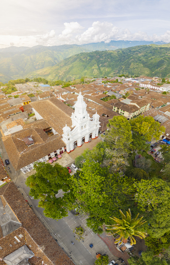 Aerial view of bright white church steeple piercing the skyline above terracotta rooftops nestled amidst lush greenery and distant mountains Salamina Caldas Colombia.