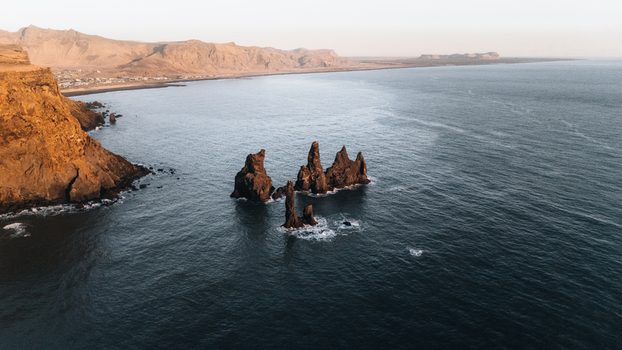 Aerial view of stark basalt sea stacks puncturing the navy blue sea at Reynisfjara beach contrasted against the golden cliffs Reynisdrangar Iceland.