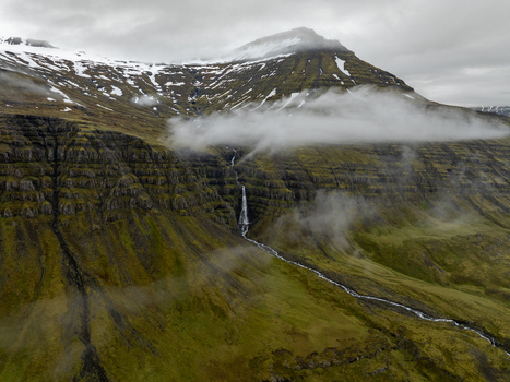 Aerial view of a waterfall cascading down a steep mossy rocky mountain slope with snow capped peaks and low hanging clouds in the Eastern Region Iceland. Aerial view of a waterfall cascading down a steep mossy rocky mountain slope with snow capped peaks and low hanging clouds in the Eastern Region Iceland.