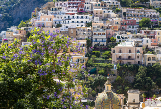 Aerial view of Positano with colorful Mediterranean houses built into the steep hillside featuring a tiled dome in the foreground and purple jacaranda flowers Positano Campania Italy. Aerial view of Positano with colorful Mediterranean houses built into the steep hillside featuring a tiled dome in the foreground and purple jacaranda flowers Positano Campania Italy.