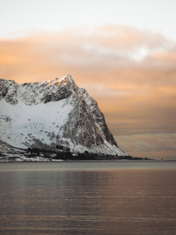 View of snow capped mountain rises majestically from the tranquil waters under a pastel colored sky a stunning contrast of cold and warm hues Indregård Troms Norway. View of snow capped mountain rises majestically from the tranquil waters under a pastel colored sky a stunning contrast of cold and warm hues Indregård Troms Norway.