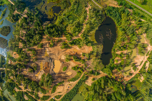 Aerial view of a sand and gravel extraction site surrounded by dense forest with muddy pools of water and winding dirt paths in Latvia. Aerial view of a sand and gravel extraction site surrounded by dense forest with muddy pools of water and winding dirt paths in Latvia.