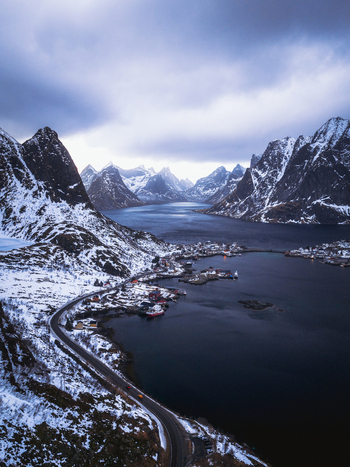 Aerial view of a winding road hugging the snow dusted coastline amidst towering mountains reflecting in the serene waters of Reinebringen Nordland Norway.