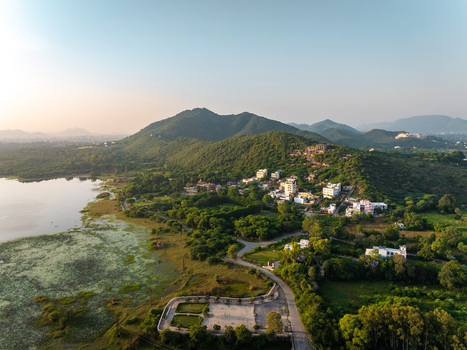 Aerial view of a serene lake nestled beside lush green hills and a small hillside settlement with a winding road and park area in Udaipur Rajasthan India.