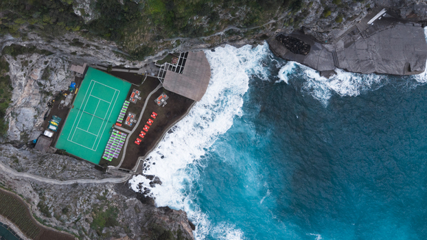 Aerial view of a vibrant green tennis court perched atop rugged cliffs where crashing turquoise waves meet the rocky shore Laurito Campania Italy. Aerial view of a vibrant green tennis court perched atop rugged cliffs where crashing turquoise waves meet the rocky shore Laurito Campania Italy.