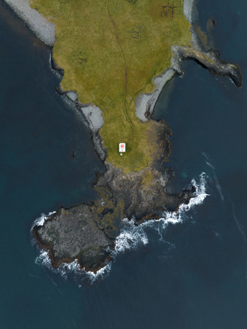 Aerial view of the rugged coastline where the verdant land meets the dark turbulent sea with a small structure standing watch K&aacute;lfshamarsv&iacute;k H&uacute;nabygg&eth; Iceland.