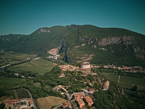 Aerial view of a historic stone fortress on a lush green hill overlooking vineyards and a winding river valley with mountains in the background Italy.