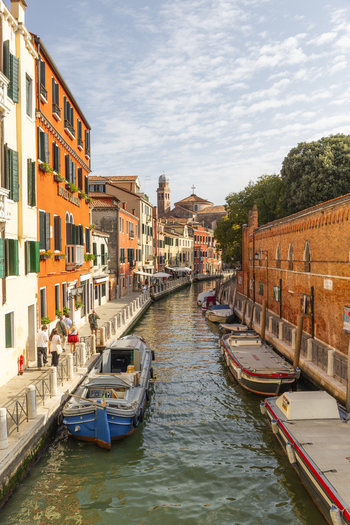 View of a narrow canal in Venice lined with colorful historic buildings moored boats and people walking along the stone embankment in Venice Veneto Italy. View of a narrow canal in Venice lined with colorful historic buildings moored boats and people walking along the stone embankment in Venice Veneto Italy.
