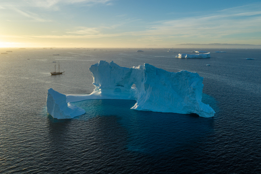 Aerial view of a large arch iceberg floating in the ocean with a sailing ship nearby during golden hour in Greenland. Aerial view of a large arch iceberg floating in the ocean with a sailing ship nearby during golden hour in Greenland.