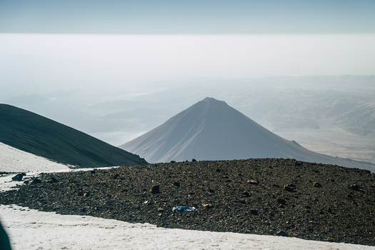 View of Mount Ararat rising above the surrounding landscape with rocky terrain and patches of snow in the foreground under a hazy sky in Turkey.