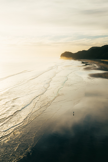 Aerial view of Piha Beach at sunset with gentle waves washing over the wet sand and a person walking along the shoreline in Auckland New Zealand. Aerial view of Piha Beach at sunset with gentle waves washing over the wet sand and a person walking along the shoreline in Auckland New Zealand.