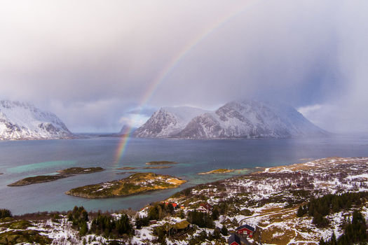 Aerial view of Lofoten with a vibrant rainbow arching over snowy mountains calm fjord waters and small islands in Nordland Norway.