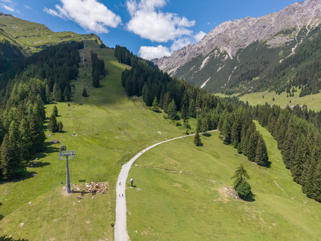 Aerial view of a mountain cable car line and a gravel path winding through lush green alpine meadows with pine forests and rocky peaks in Imst Tyrol Austria.