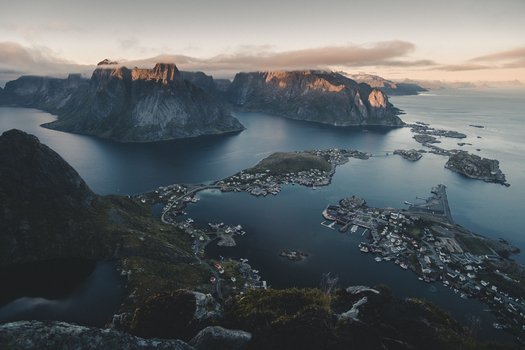 View of the village of Reine and surrounding fjords and mountains from the peak of Reinebringen with calm water rocky terrain and soft golden hour light in Moskenes Nordland Norway.
