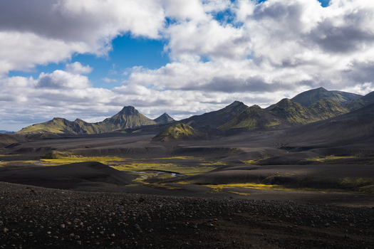 View of stark dark volcanic landscape punctuated by patches of vibrant green moss and distant craggy mountains under a sky of dramatic clouds Skaftárhreppur Iceland. View of stark dark volcanic landscape punctuated by patches of vibrant green moss and distant craggy mountains under a sky of dramatic clouds Skaftárhreppur Iceland.
