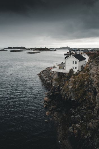 View of the coastal village of Henningsvaer with a white house perched on rocky cliffs overlooking the sea under a dramatic cloudy sky in Henningsvaer Nordland Norway. View of the coastal village of Henningsvaer with a white house perched on rocky cliffs overlooking the sea under a dramatic cloudy sky in Henningsvaer Nordland Norway.