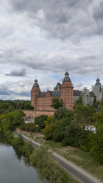 Aerial view of Schloss Johannisburg a historic red sandstone castle situated along the Main river surrounded by lush trees and greenery under a cloudy sky in Aschaffenburg Bayern Germany.