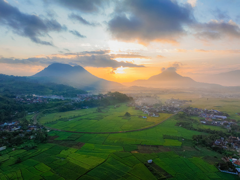 Aerial view of lush green rice fields surrounding a village nestled between mountains during a golden sunrise in Kadungora West Java Indonesia. Aerial view of lush green rice fields surrounding a village nestled between mountains during a golden sunrise in Kadungora West Java Indonesia.