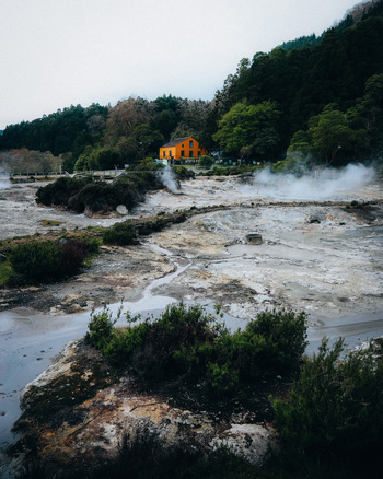 View of steaming vents and volcanic landscape contrasting with the lush green forest and a vibrant yellow house Furnas Azores Portugal.