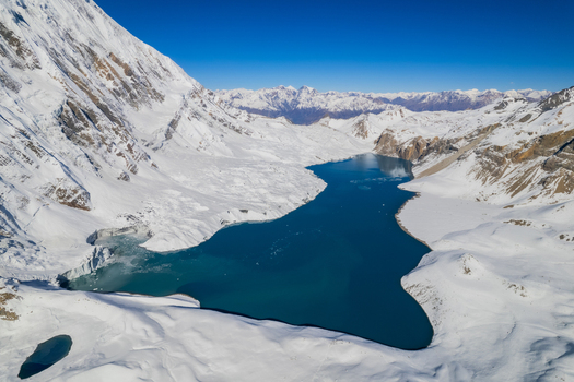 Aerial view of a deep blue glacial lake nestled among snow covered Himalayan peaks under a clear blue sky in Nepal.