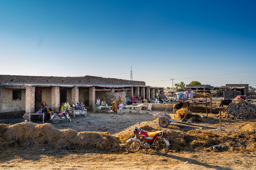 View of a rural settlement with local residents sitting outside a brick building motorcycles livestock and hay piles in Jacobabad Sindh Pakistan.