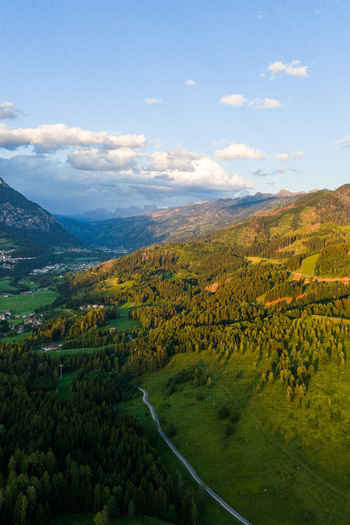 Aerial view of Fiemme Valley featuring lush green forests winding mountain roads and dramatic peaks illuminated by golden hour light in Trentino Alto Adige Italy.