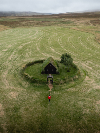 Aerial view of a solitary figure approaching the turf roofed Hofs&oacute;s church amidst a verdant field a haven of tranquility under a cloudy sky Hofs&oacute;s Skagafj&ouml;r&eth;ur Iceland.