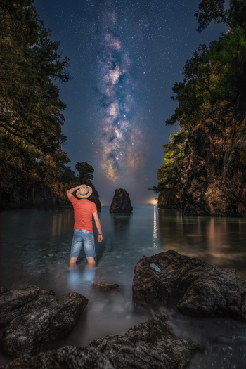 View of a man in a straw hat standing in the sea looking at the Milky Way galaxy between rocky cliffs at night in Lerici Liguria Italy.