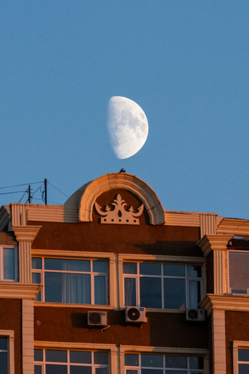 View of a building rooftop with a crown like architectural detail illuminated by warm sunset light under a clear blue sky with a half moon in Aktau Mangystau Region Kazakhstan.