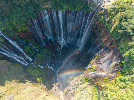Aerial view of Tumpak Sewu Waterfall featuring cascading streams over a lush green cliffside with a visible rainbow in the mist in Kecamatan Pronojiwo Jawa Timur Indonesia.