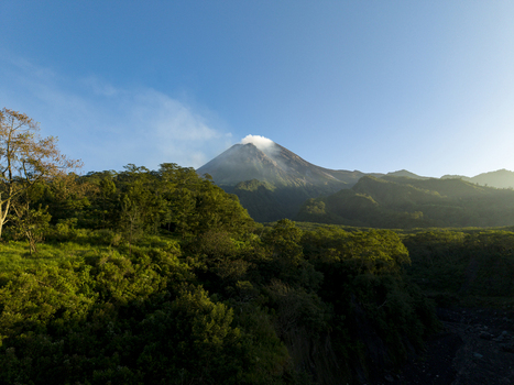 Aerial view of Mount Merapi volcano with smoke rising from the crater above lush green forests under a clear blue sky in Kecamatan Pakem Daerah Istimewa Yogya Indonesia. Aerial view of Mount Merapi volcano with smoke rising from the crater above lush green forests under a clear blue sky in Kecamatan Pakem Daerah Istimewa Yogya Indonesia.