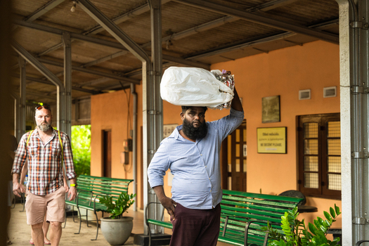 Sri Lanka 06 February 2026 View of a man carrying a large white sack on his head while walking past green benches in a covered corridor.
