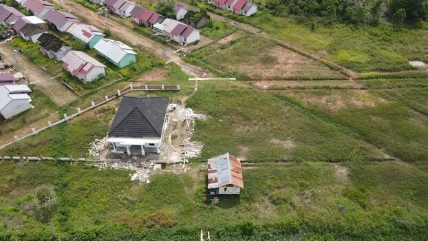 Aerial view of a residential construction site featuring a new house with a dark roof and a small shed surrounded by grassy fields and rows of houses in Palembang South Sumatra Indonesia. Aerial view of a residential construction site featuring a new house with a dark roof and a small shed surrounded by grassy fields and rows of houses in Palembang South Sumatra Indonesia.