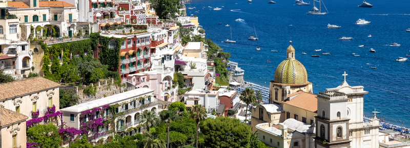 Aerial view of the colorful buildings of Positano featuring the majolica tiled dome of the Church of Santa Maria Assunta overlooking the blue sea with boats in Positano Campania Italy. Aerial view of the colorful buildings of Positano featuring the majolica tiled dome of the Church of Santa Maria Assunta overlooking the blue sea with boats in Positano Campania Italy.